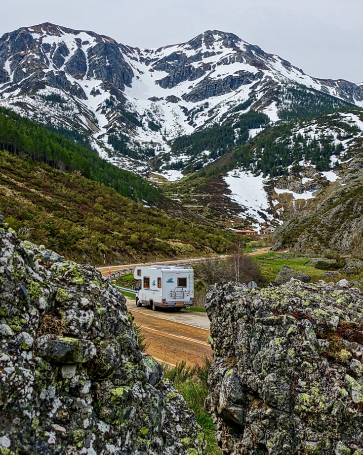 A campervan driving through a valley surrounded by snow capped mountains