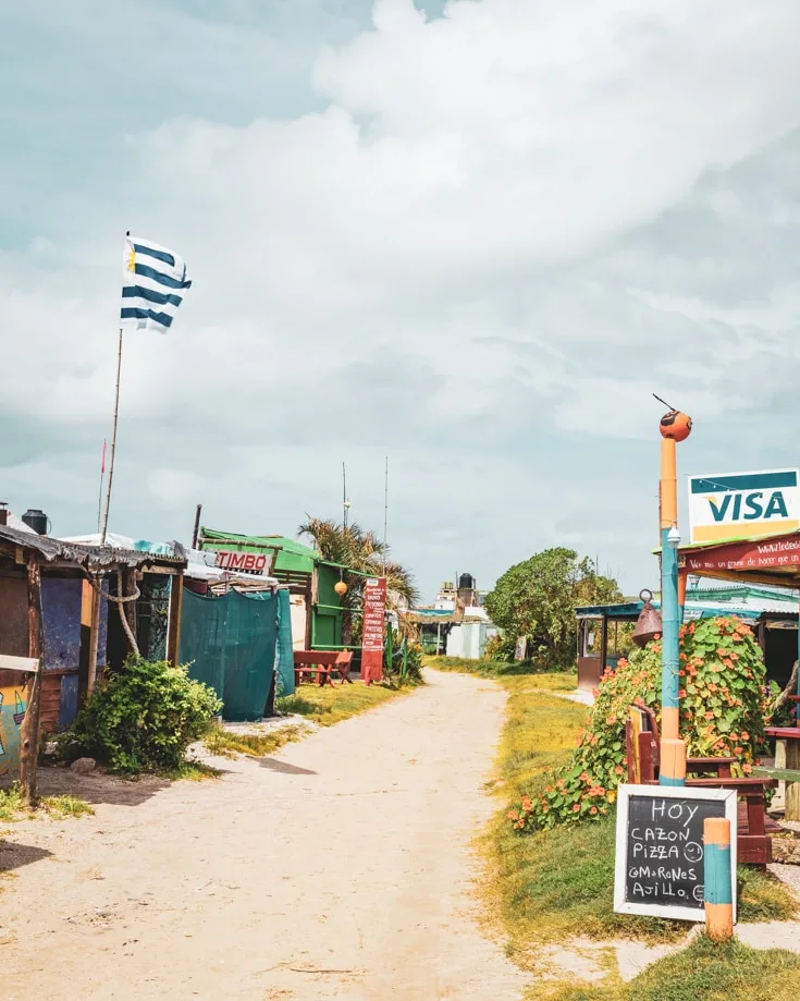 Sandy tracks in Cabo Polonio with the Uruguayan flag flying and a visa sign outside a bar
