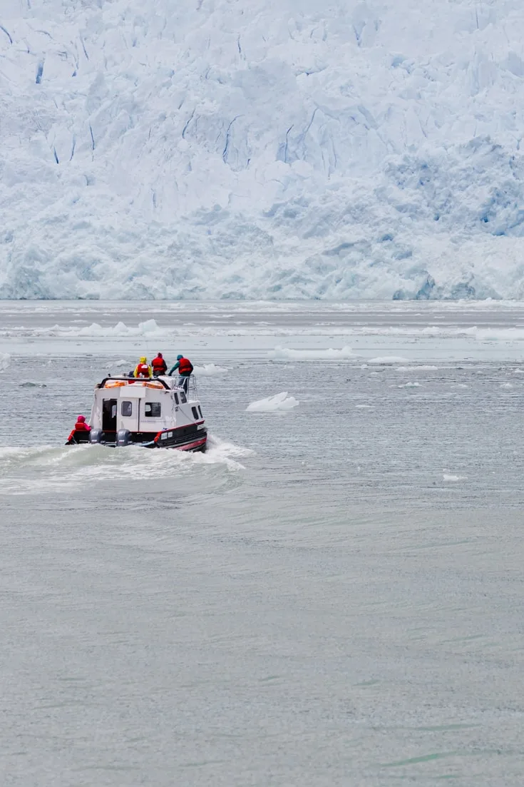 A small tour boat with a a few passengers sails towards the face of a glacier through brash ice