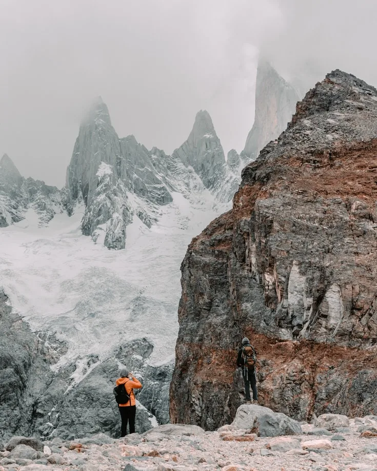 A couple hiking in Patagonia and looking up at a glacier and mountain peaks shrouded in cloud