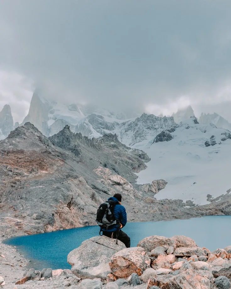a hiker sitting on a boulder at the top of Mount Fitz Roy hike with a lake in the foreground