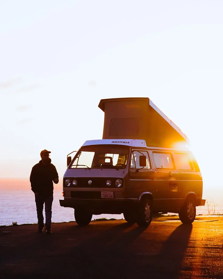 A man standing beside a VW kombi pop top campervan at sunset