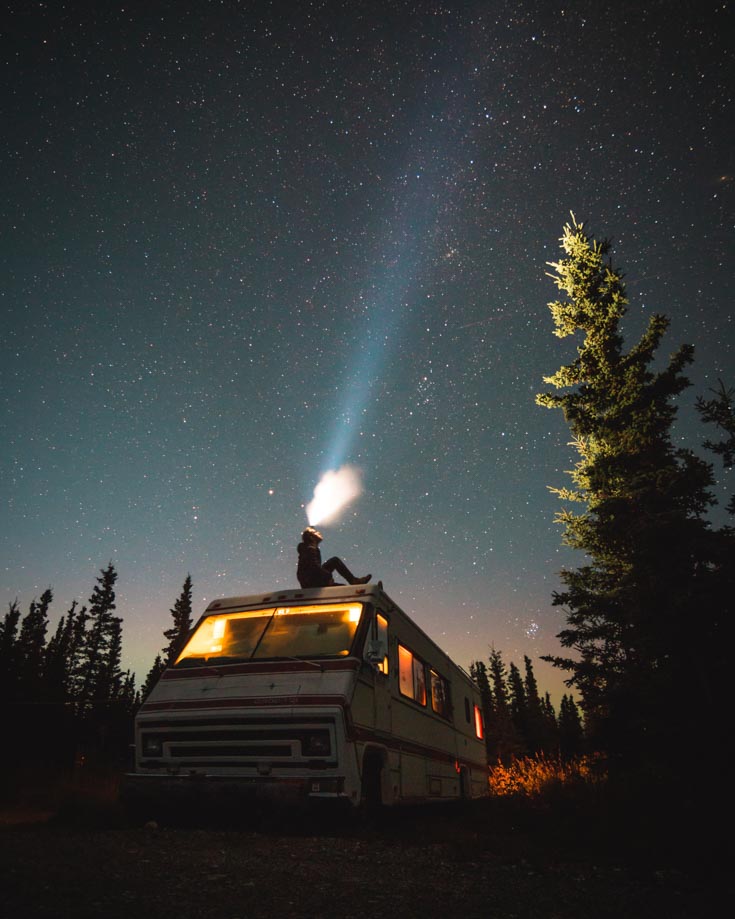 a person sitting on the roof of a camper van at night shining a bright torch towards the milky way