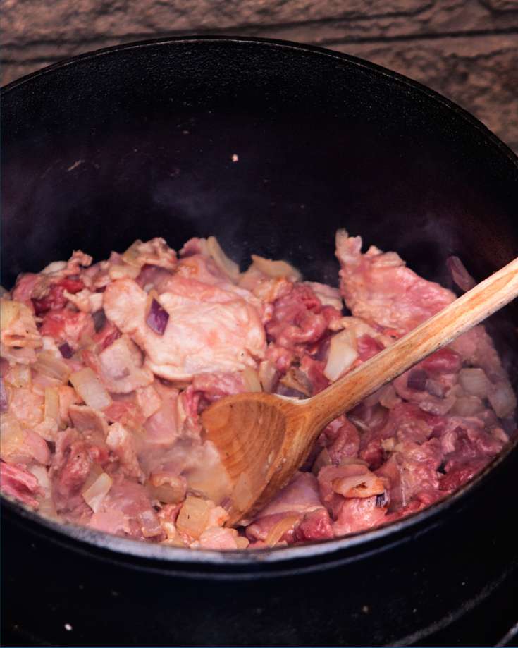 A campfire-side view of a one-pot meal being prepared, with ingredients like chopped vegetables and spices laid out nearby.